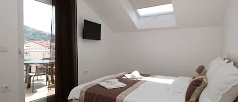 Une chambre avec un lit, une télévision, un balcon avec une table et des chaises, et une vue sur les bâtiments environnants.