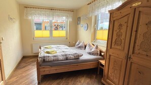 Quarto com cama de madeira, duas janelas com persianas amarelas, radiador e guarda-roupa de madeira.