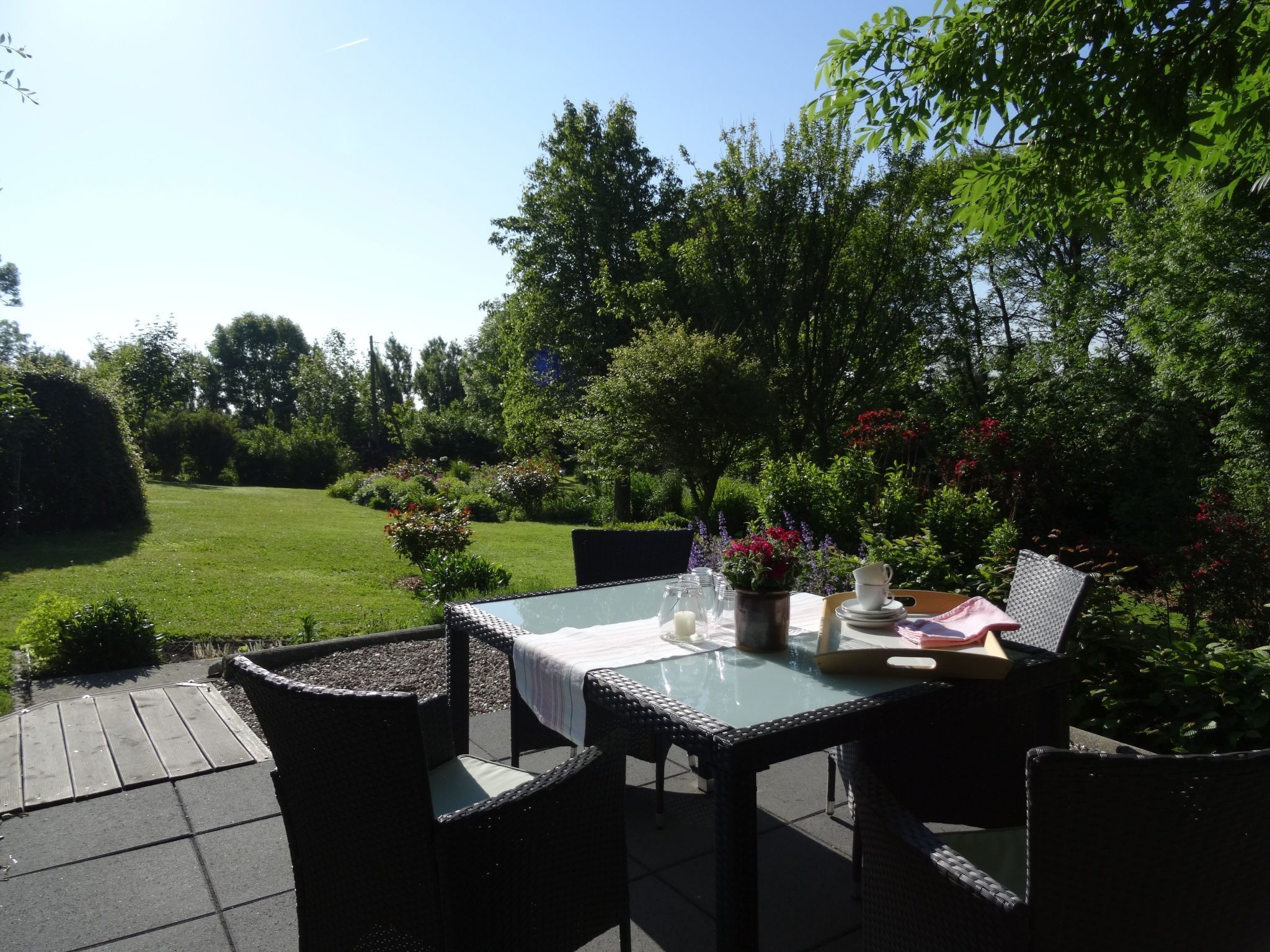 Une terrasse extérieure avec une table en verre et des chaises en osier, entourée d’une végétation luxuriante et d’une pelouse bien entretenue.