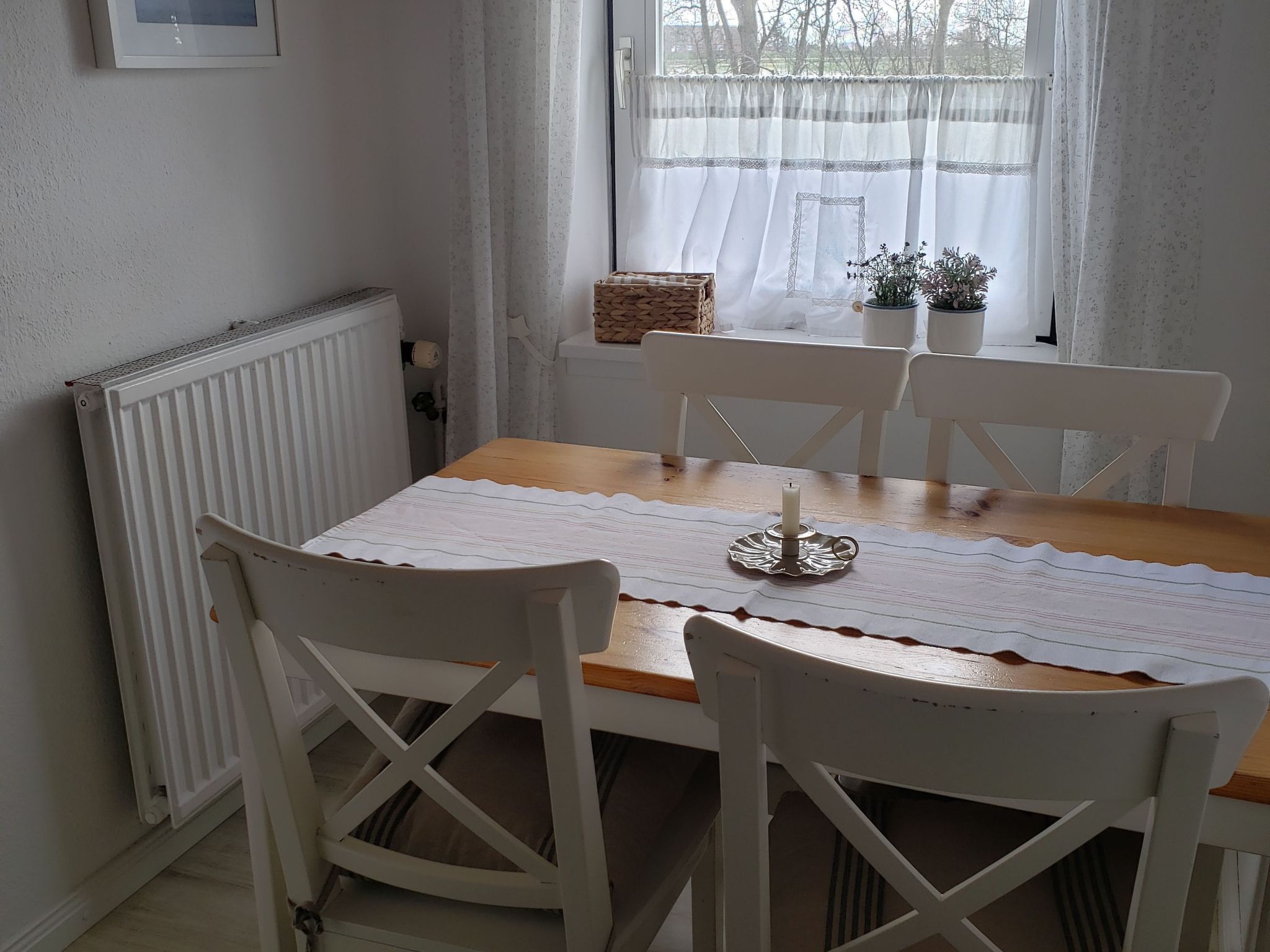 A dining area with a wooden table, white chairs, a white tablecloth, a candle holder, and a window with sheer curtains.