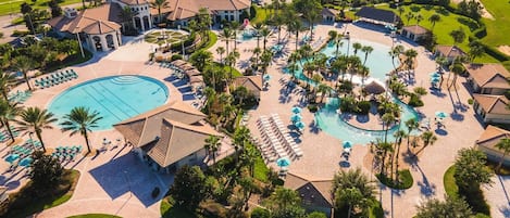 An aerial view of a resort with a large central pool, multiple smaller pools, and a well-maintained landscape with palm trees and pathways.