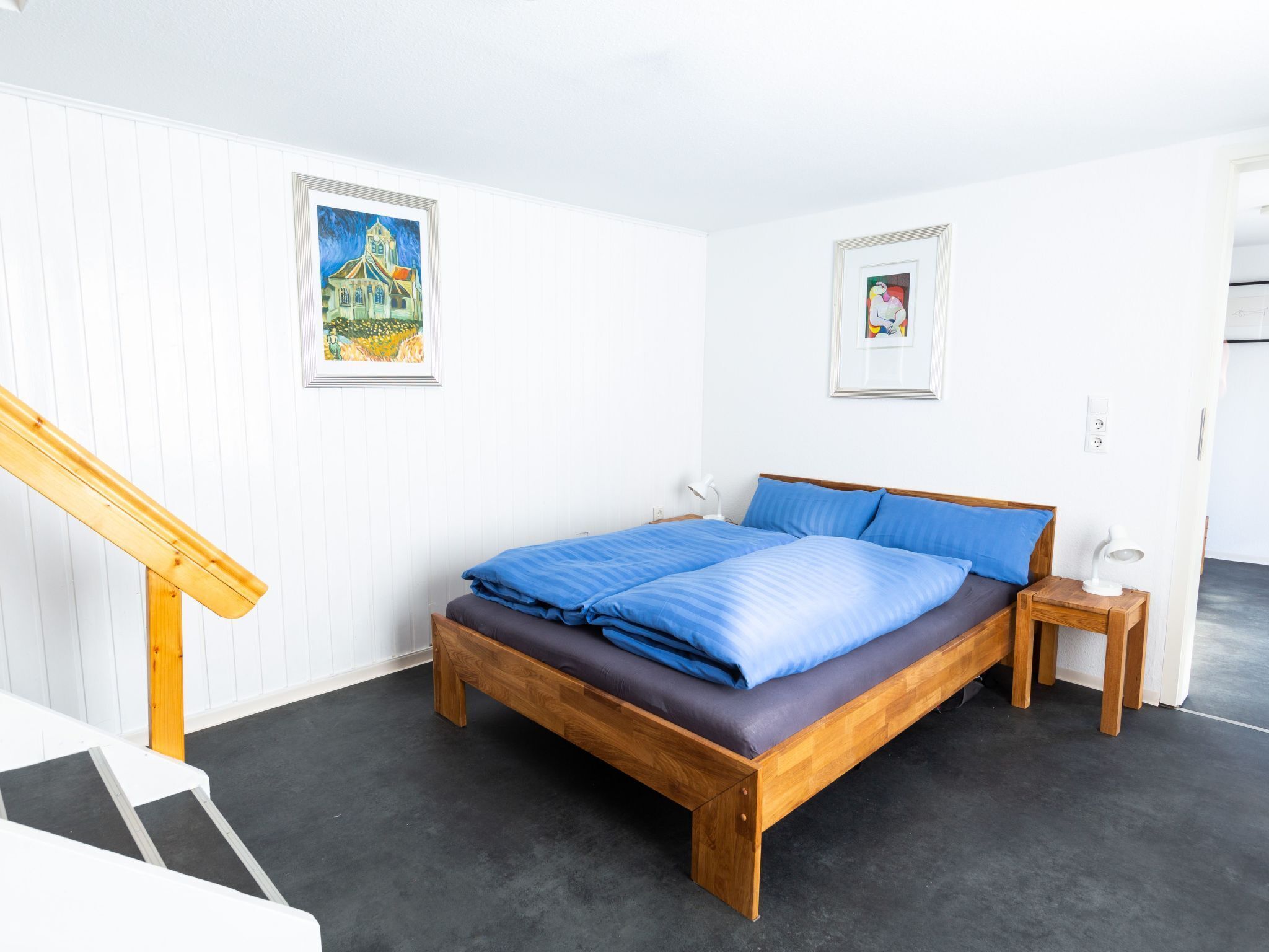 A bedroom with a wooden bed, a nightstand, a desk, and a chair. There are two framed pictures on the wall.