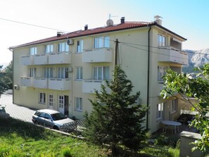 Un immeuble résidentiel à plusieurs étages, doté de balcons, avec une voiture garée devant et un paysage montagneux en arrière-plan.