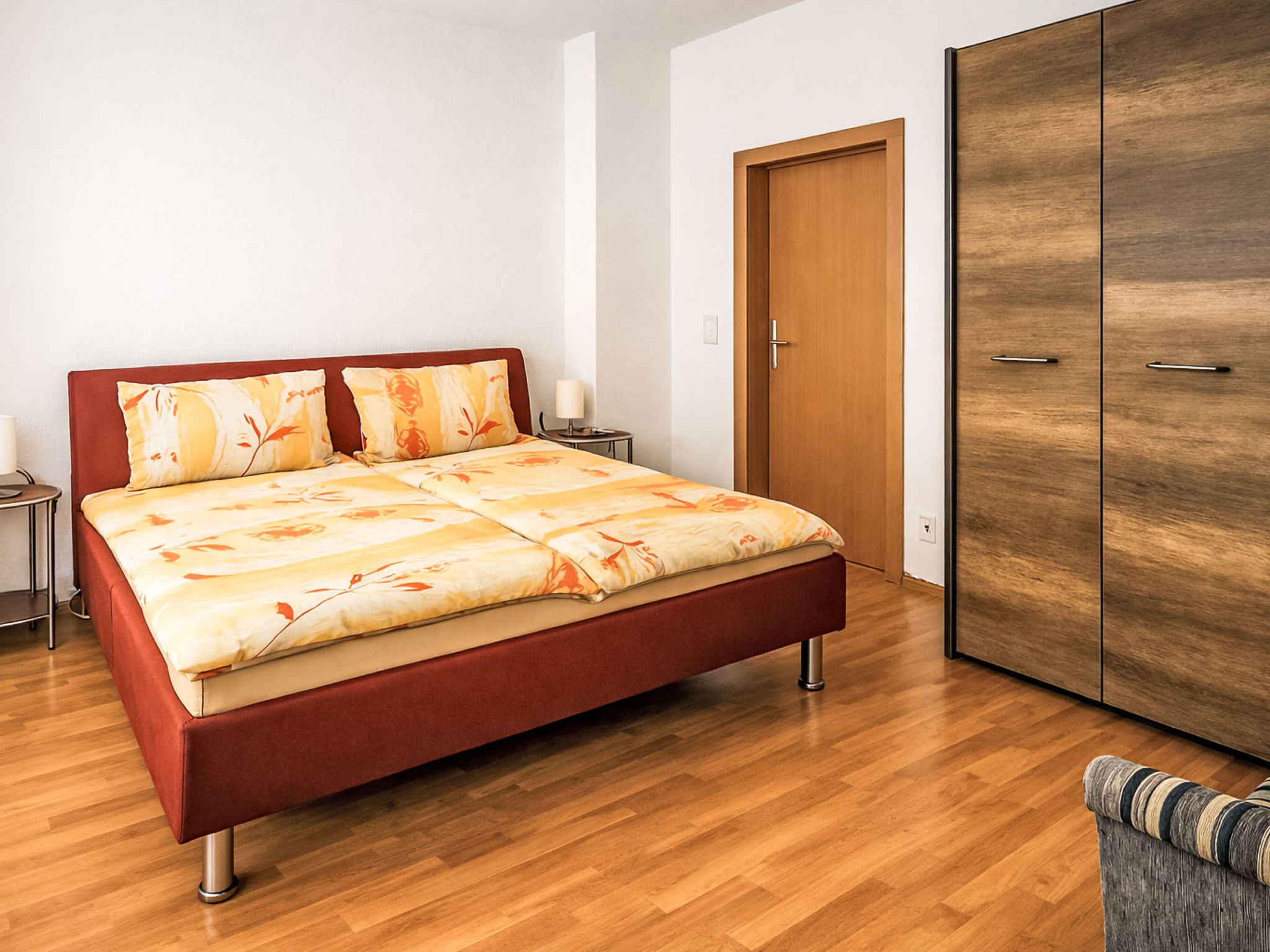 A bed with a floral bedspread, a wooden wardrobe, and a nightstand with a lamp.