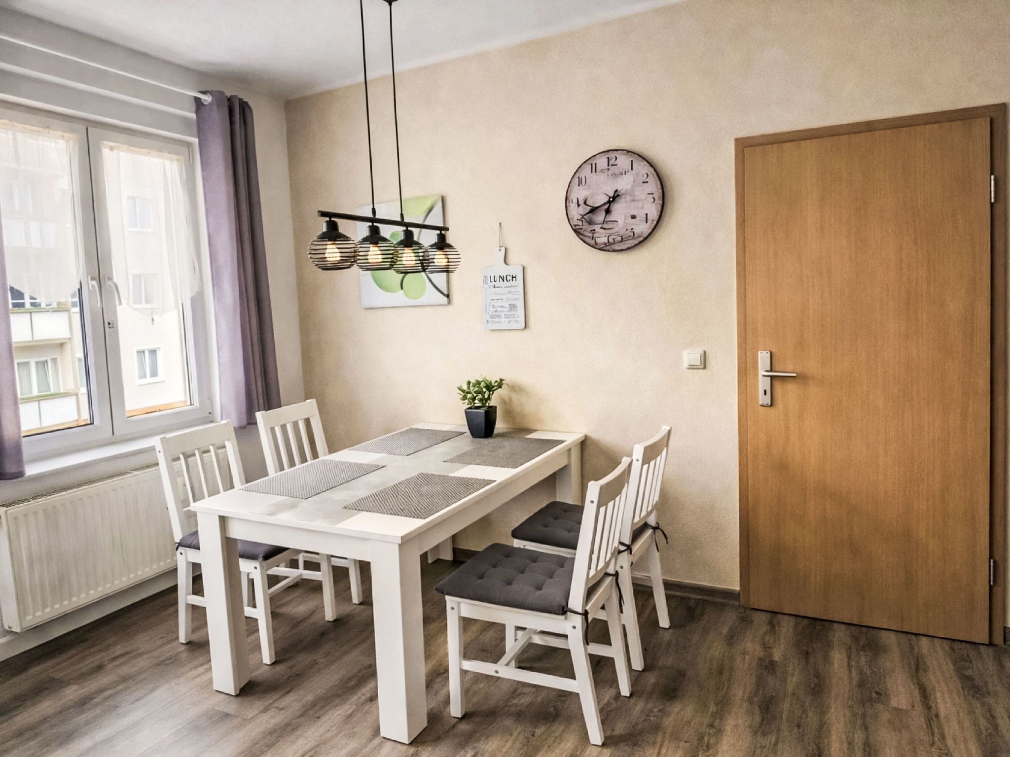 Ein Essbereich mit einem weißen Tisch und Stühlen, einer Uhr an der Wand und einer Tür rechts.