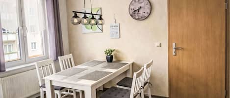 Ein Essbereich mit einem weißen Tisch und Stühlen, einer Uhr an der Wand und einer Tür rechts.