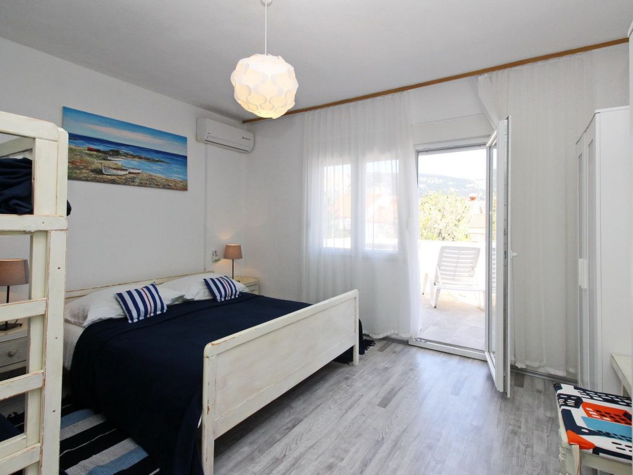 A bedroom with a bed, a nightstand, a lamp, a painting, and a view of the outdoors.