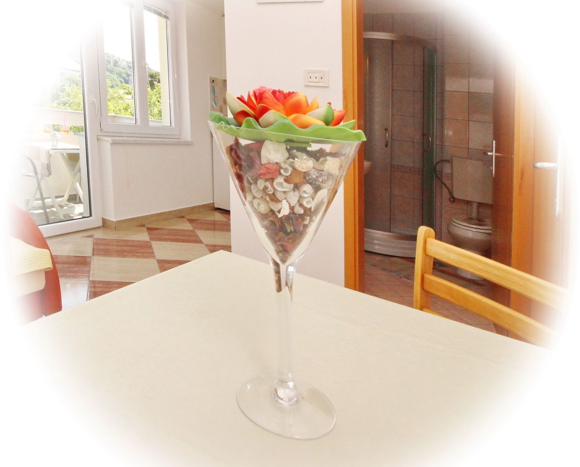 En genomskinlig glasvas med en dekorativ blomsteruppsättning på ett bord i ett rum med ett klinkergolv och ett synligt område för dusch.