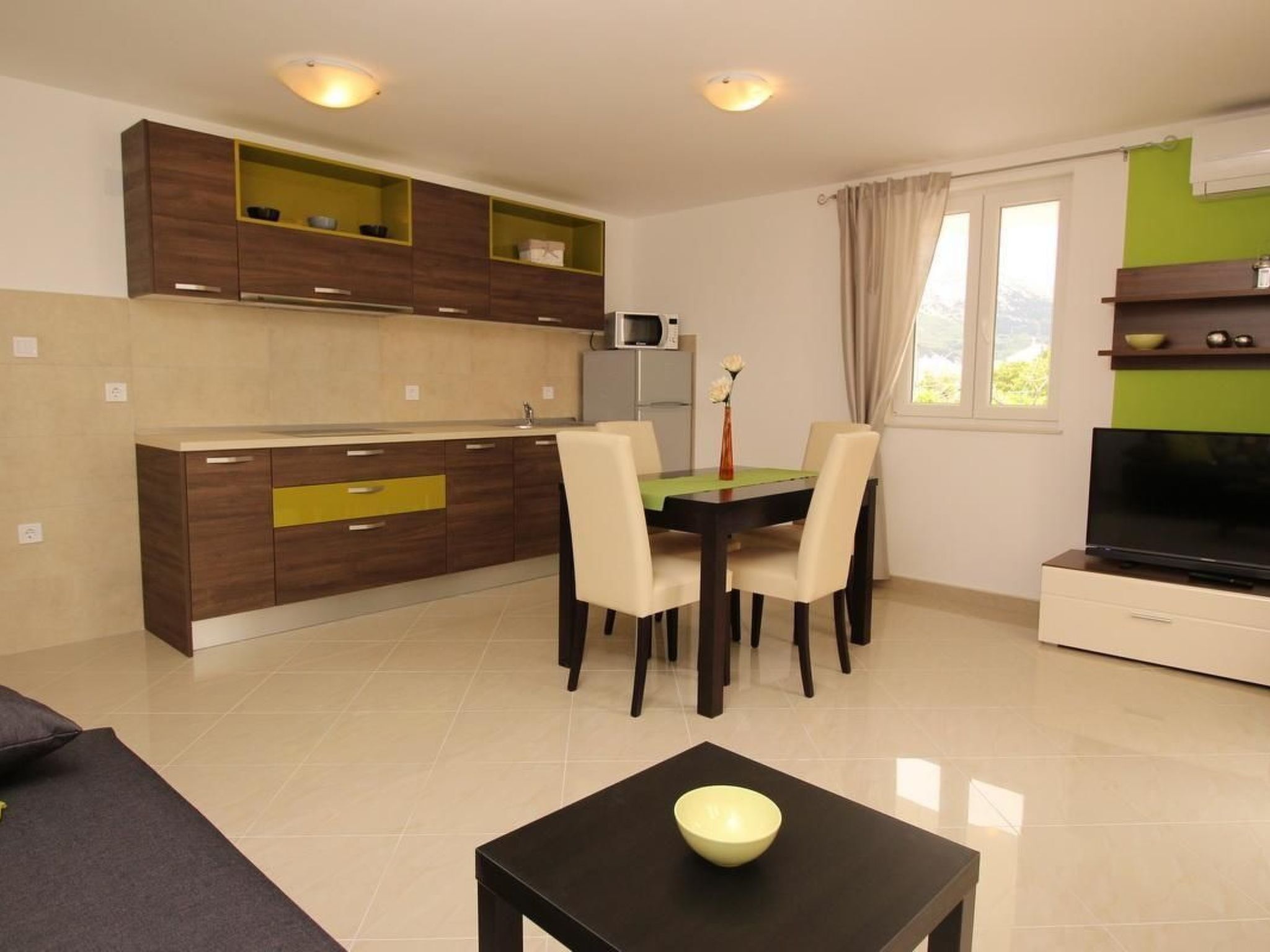 A modern kitchen with wooden cabinets, a dining table with chairs, a flat-screen TV, and a small table with a bowl.