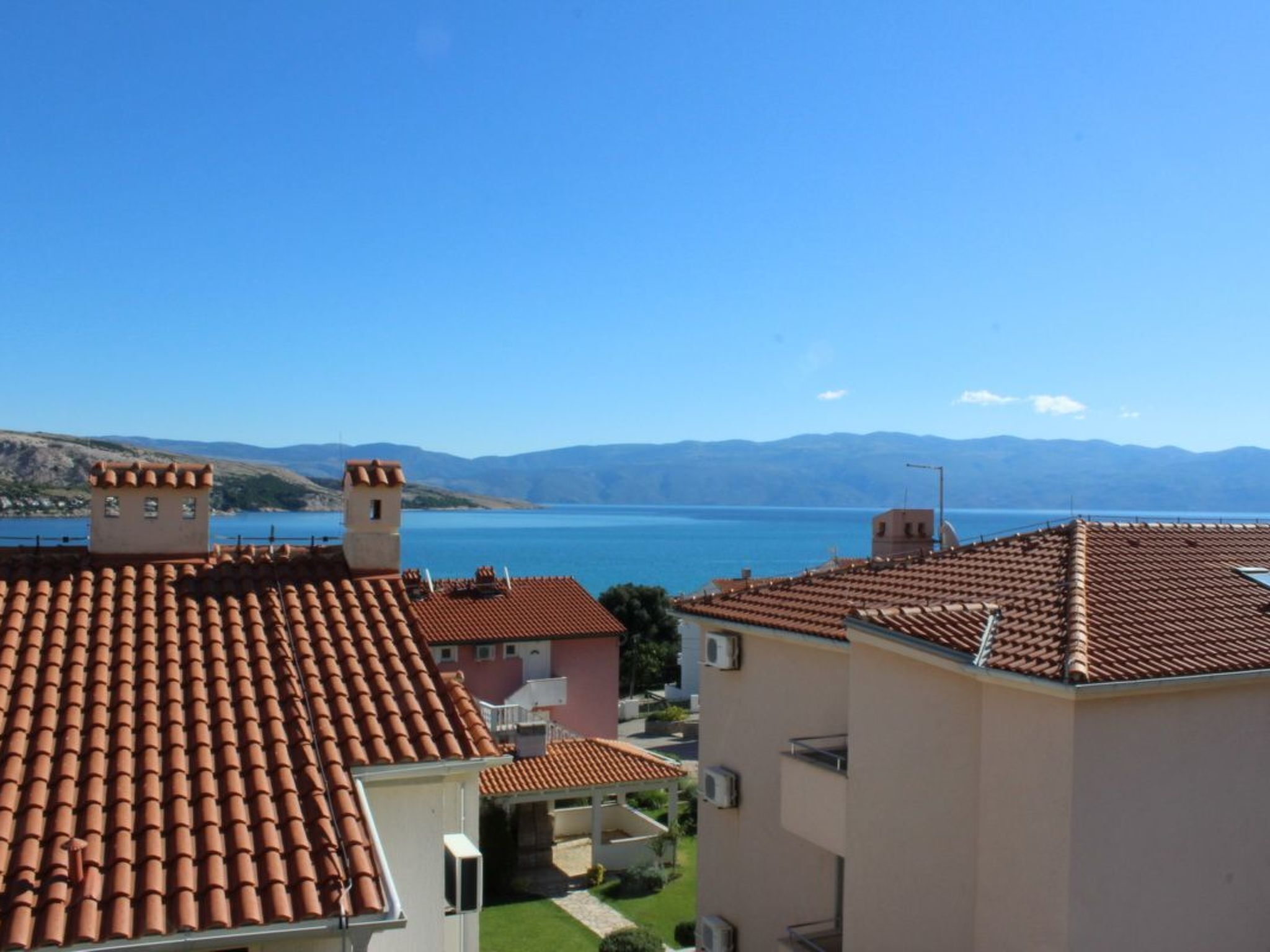 A residential area with terracotta-tiled roofs, a clear blue sky, and a view of a lake and mountains in the distance.