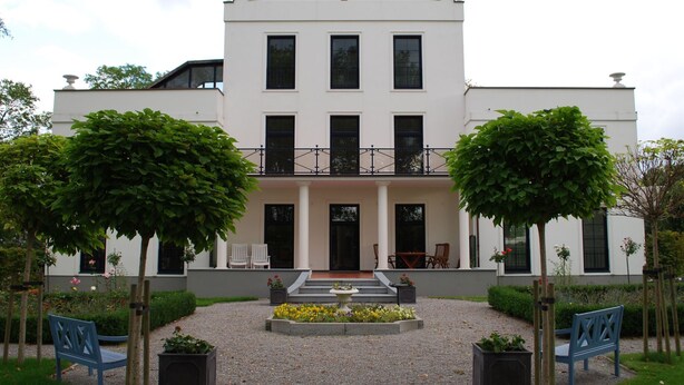 Un edificio a due piani con facciata simmetrica, ampie finestre e un ingresso centrale con gradini che conducono a una porta.