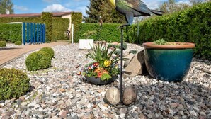 A birdhouse with a bird perched on it, surrounded by a garden with gravel, plants, and a blue fence in the background. - Ferienhaus Claire am Klostersee Urlaub mit Hund by Interhome (Dargun)