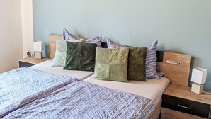 Ein Bett mit gemustertem Überwurf, ein Kopfteil mit vielen Kissen, ein Nachttisch mit Lampe und ein an der Wand befestigtes Kunstwerk, das eine Welle zeigt.