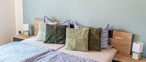 Ein Bett mit gemustertem Überwurf, ein Kopfteil mit vielen Kissen, ein Nachttisch mit Lampe und ein an der Wand befestigtes Kunstwerk, das eine Welle zeigt.