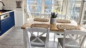 A dining area with a wooden table, white chairs, and woven placemats. A potted plant is on the table, and there is a view of the outdoors through large windows. - Strandhaus am Wasser by Interhome (Altwarp)