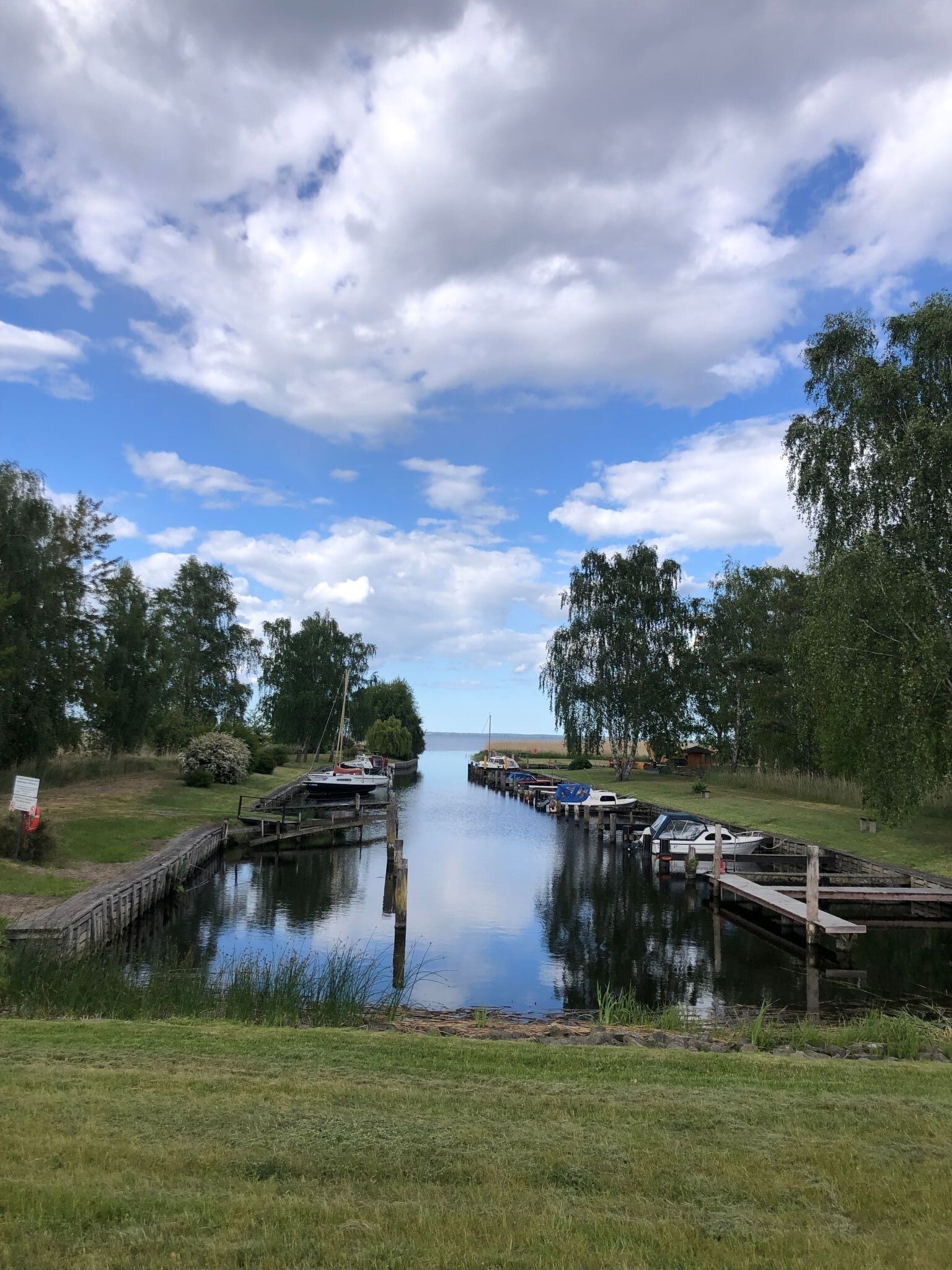 A calm waterway with boats docked on wooden piers, surrounded by grassy banks and trees.