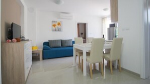 Un salón moderno y bien iluminado con un sofá azul, una mesa de comedor con sillas y una cocina al fondo.