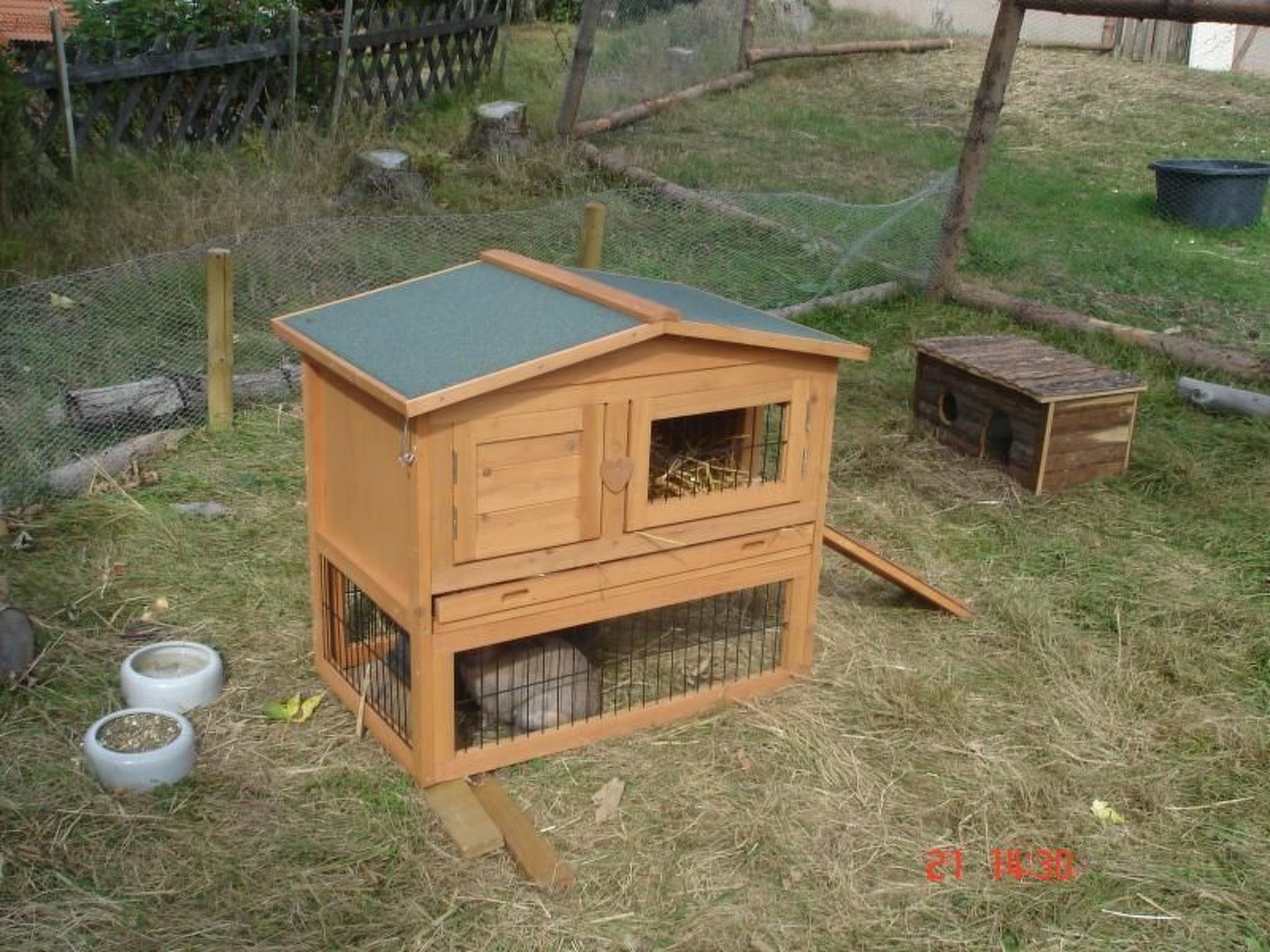 Un poulailler en bois avec un toit vert, un petit abri attenant et deux gamelles posées sur le sol.