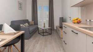 Eine moderne Wohnung mit grauem Sofa, einem kleinen Esstisch, einer Küchenzeile mit Obstschale und einem Fernseher.