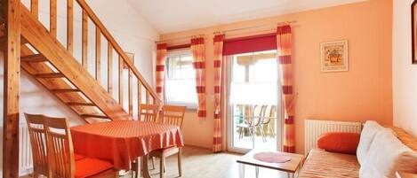 A cozy living room with a wooden staircase, a dining area with a red tablecloth, and a sofa with orange cushions.