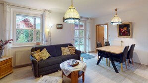 A cozy living room with a sofa, a dining table with chairs, and a small ottoman. - Christine by Interhome (Bischofswiesen)