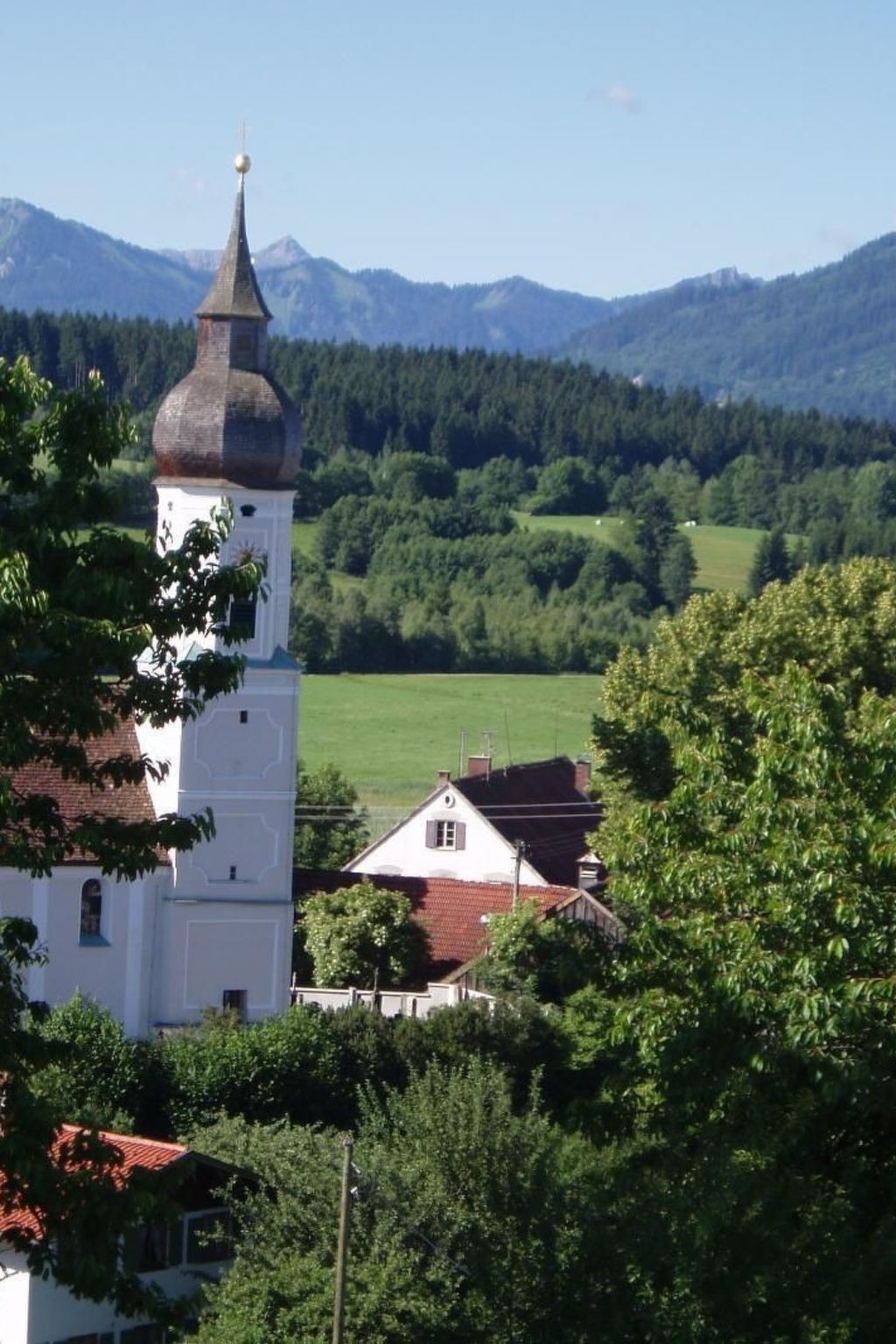 Une église blanche avec un clocher et une maison à toit rouge, nichées dans une vallée verdoyante, avec des montagnes en arrière-plan.