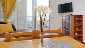 Un salon avec une table à manger en bois, des chaises, un vase avec des fleurs blanches, un téléviseur fixé au mur et un meuble en bois contenant des objets décoratifs.