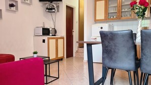 Un espacio habitable compacto con kitchenette, zona de comedor con dos sillas, televisor montado en la pared y una pequeña mesa con una planta.