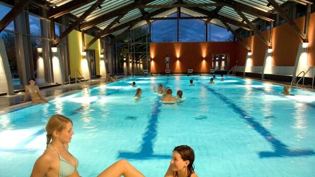 En indendørs swimmingpool med flere baner, et overdækket tag og udsigt til det frie landskab i skumringen.