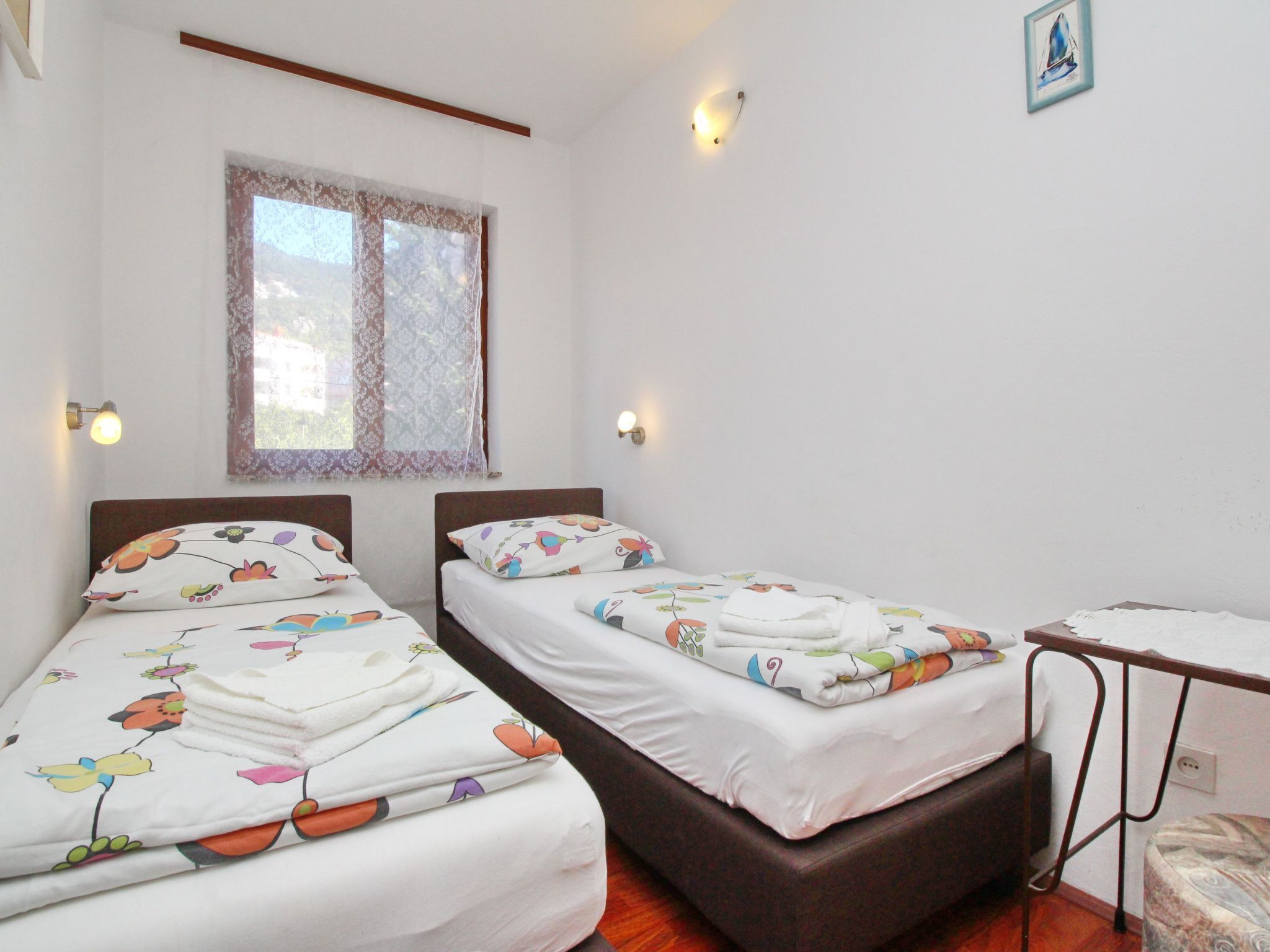 A room with two beds, each with floral-patterned bedding, a window with lace curtains, and wall-mounted lights.