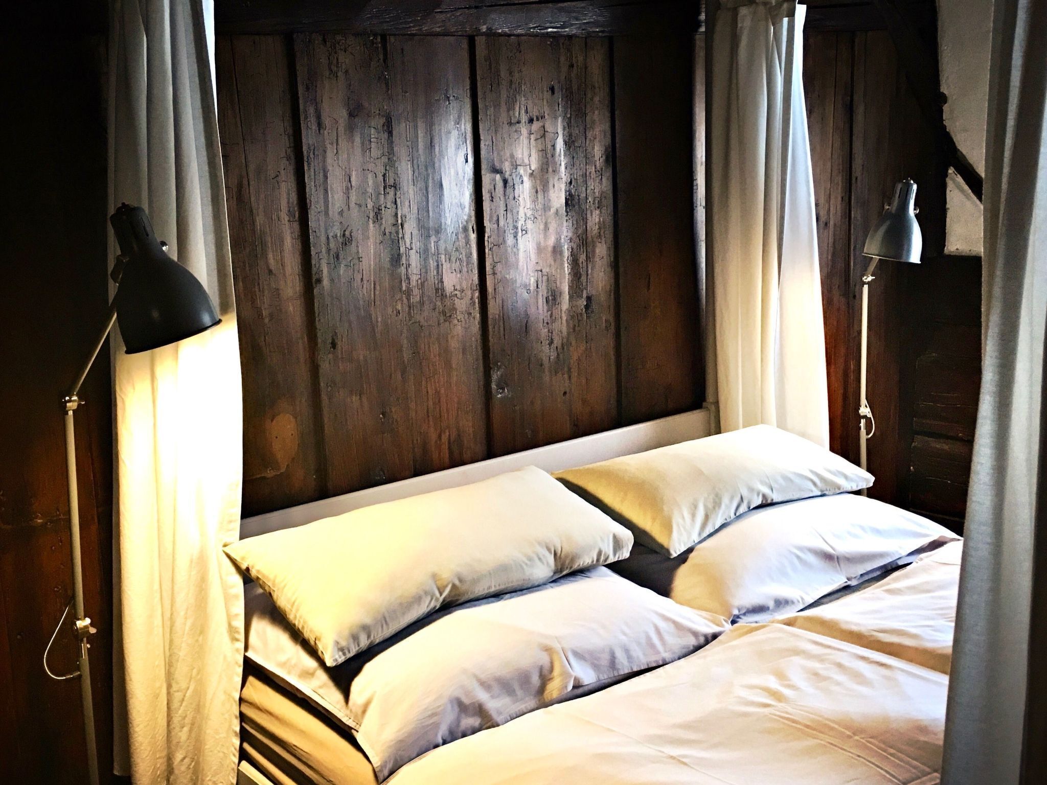 Cama com lençóis brancos num quarto com paredes de madeira e uma luminária de metal de cada lado.