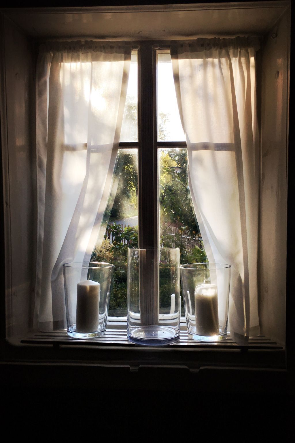 Ein Fenster mit durchsichtigen Vorhängen, einer Kerze und drei Gläsern auf einem Fensterbrett.