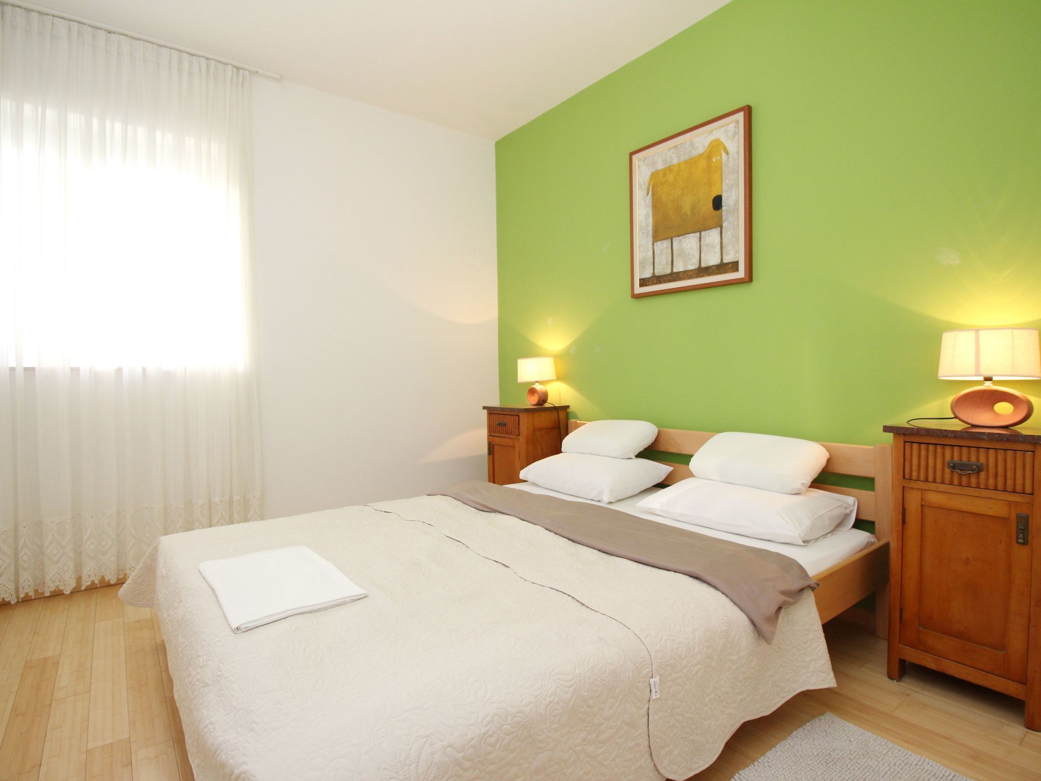 A neatly made bed with white linens, a bedside table with a lamp, and a green wall with a framed picture.