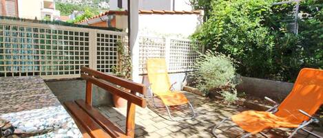 An outdoor patio with a wooden bench, a table with a pebble surface, and two orange lounge chairs.