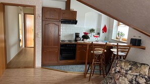 A compact kitchen with wooden cabinets, a built-in oven, a coffee maker, and a small dining area with a chair and table.