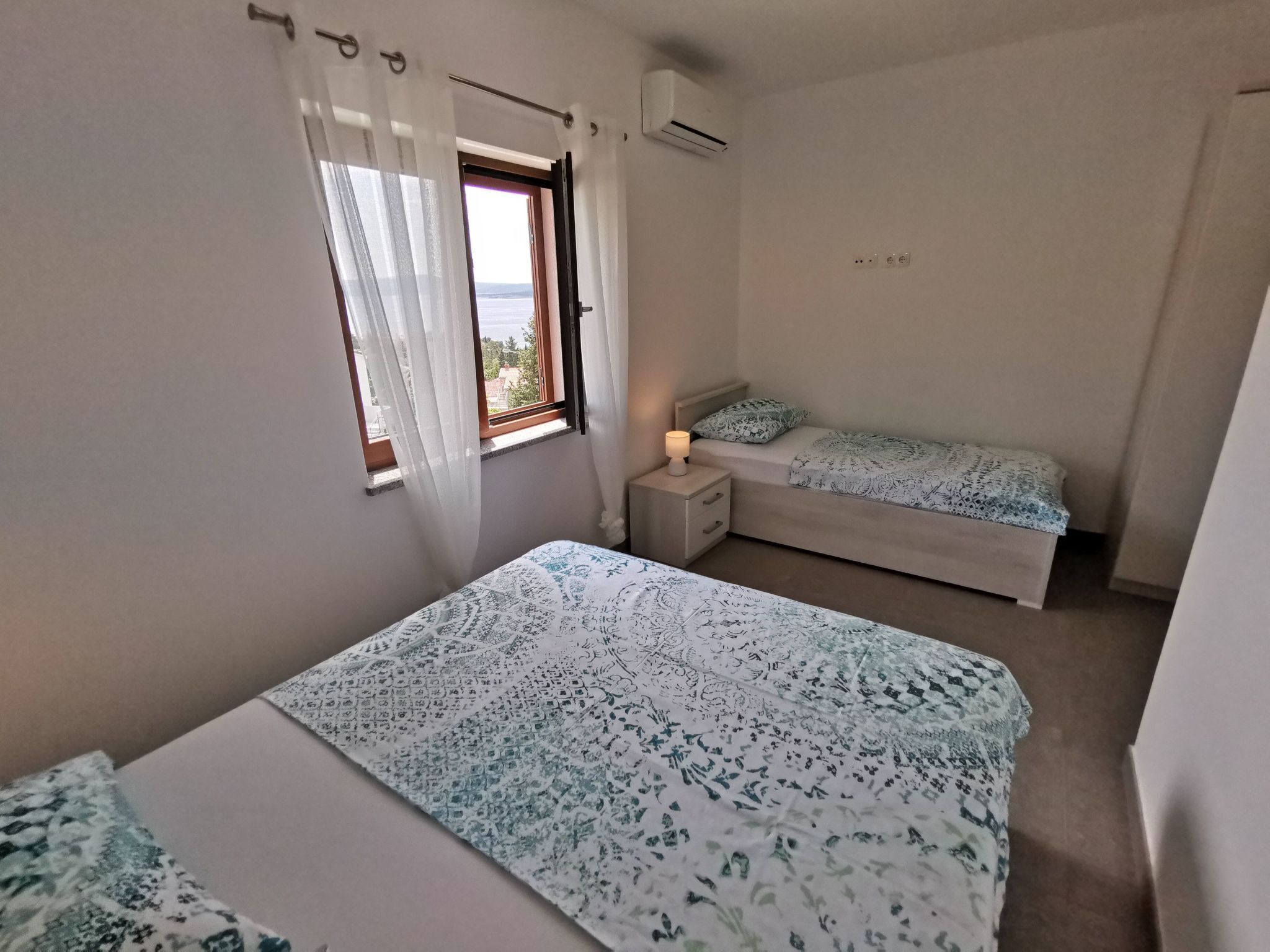 Une chambre avec un grand lit, un plus petit lit, une fenêtre avec des rideaux, un appareil de climatisation et une lampe de chevet.