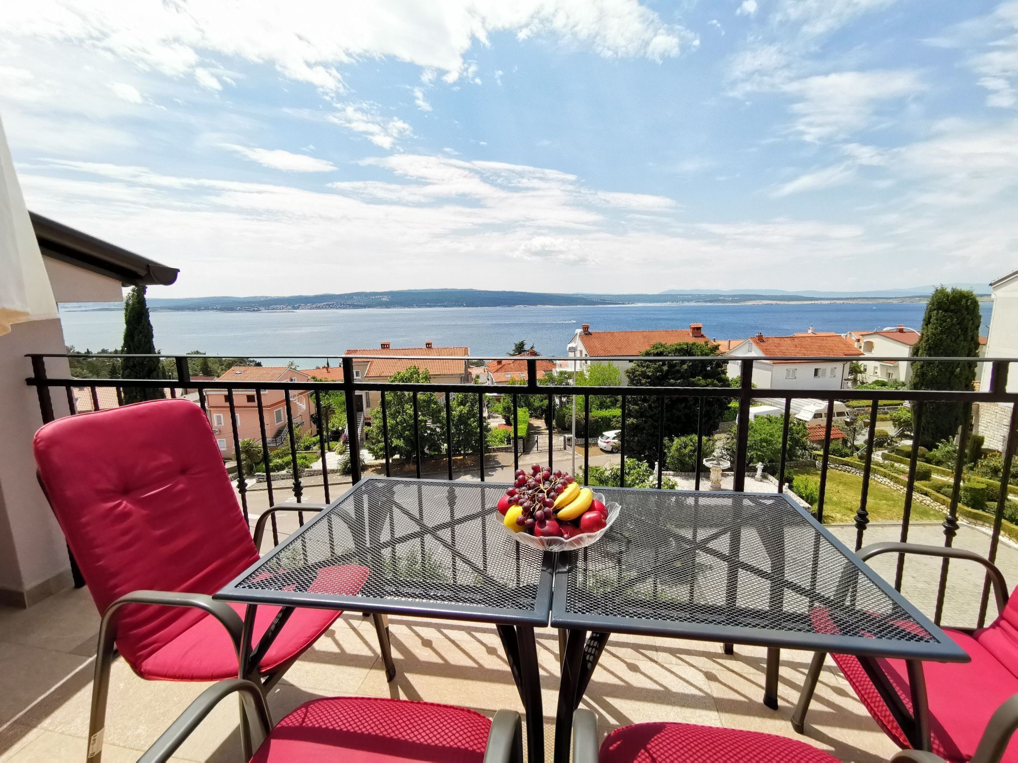 Een balkon met een tafel en stoelen, een schaal fruit en uitzicht op zee en gebouwen.