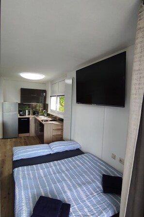 Ein kleines Zimmer mit Bett, einem an der Wand befestigten Fernseher, einer Küchenzeile und einem Fenster mit Vorhängen.