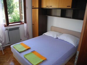 A bedroom with a bed, wooden cabinets above, a window with curtains, and a radiator below. - für 4 Personen by Interhome (Crikvenica)