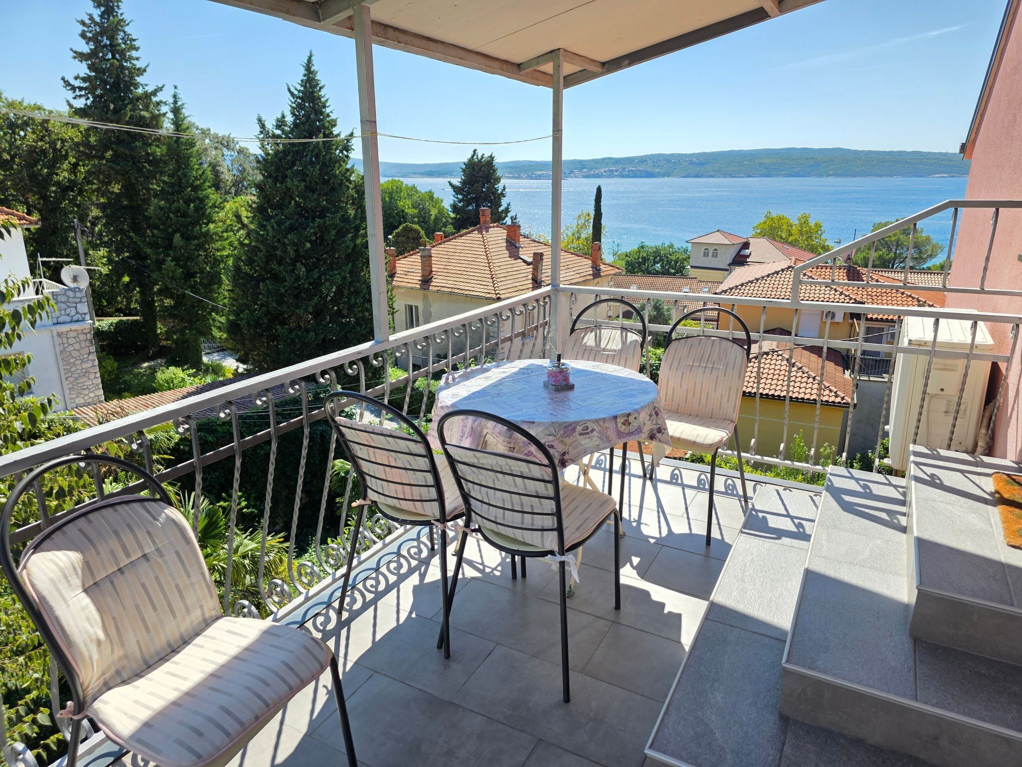 A balcony with a table and chairs overlooking a lake, surrounded by greenery and buildings.