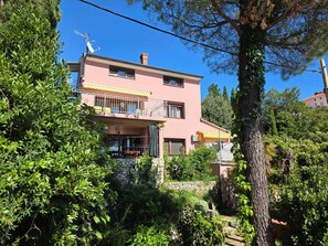 Ein zweistöckiges Gebäude mit Balkon, umgeben von üppiger Vegetation und einer Steinmauer.