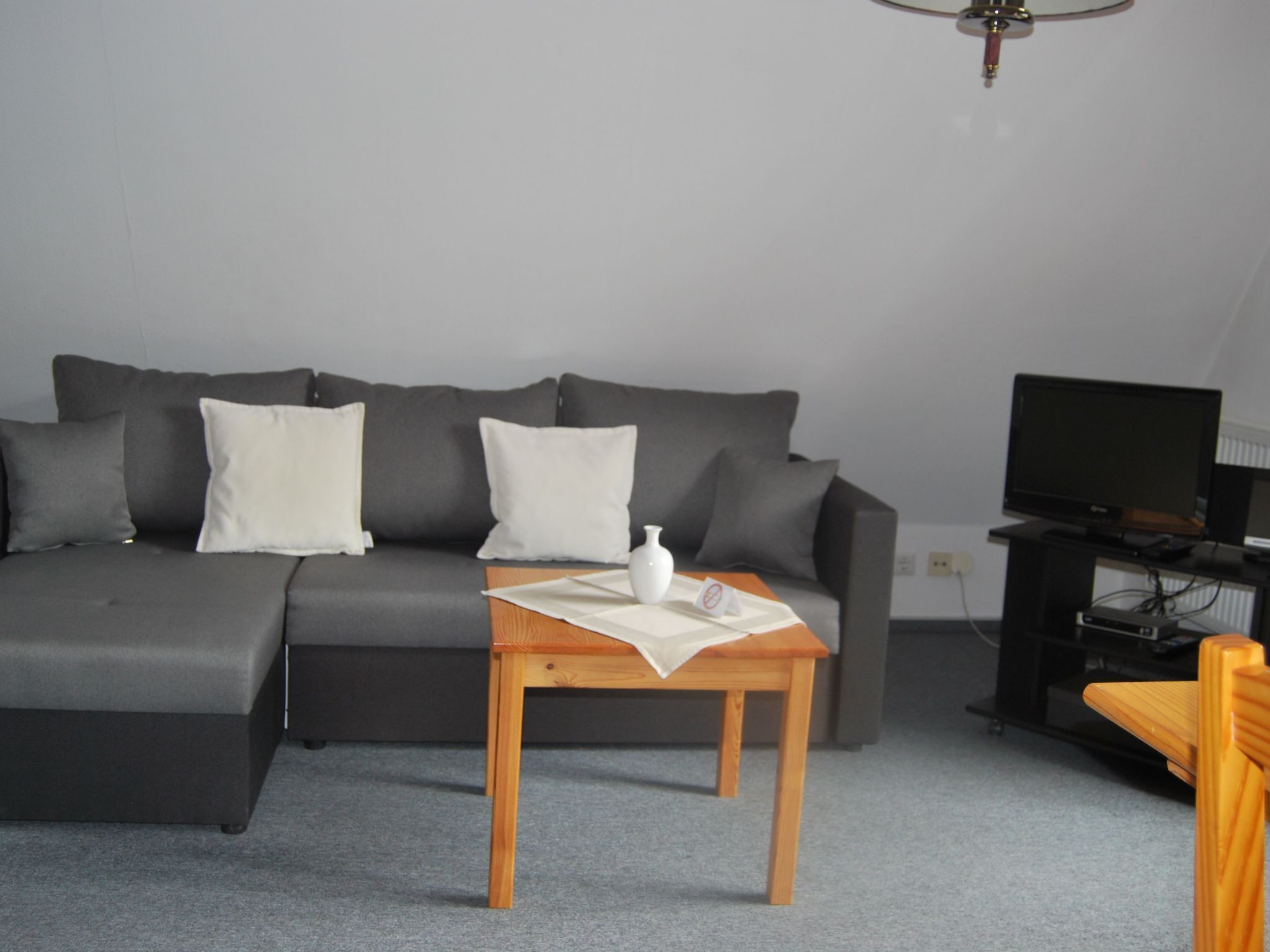 Ein modernes Wohnzimmer mit einem grauen Sofa, einem Holztisch, einem Fernseher und einem kleinen Tisch mit einer weißen Vase.