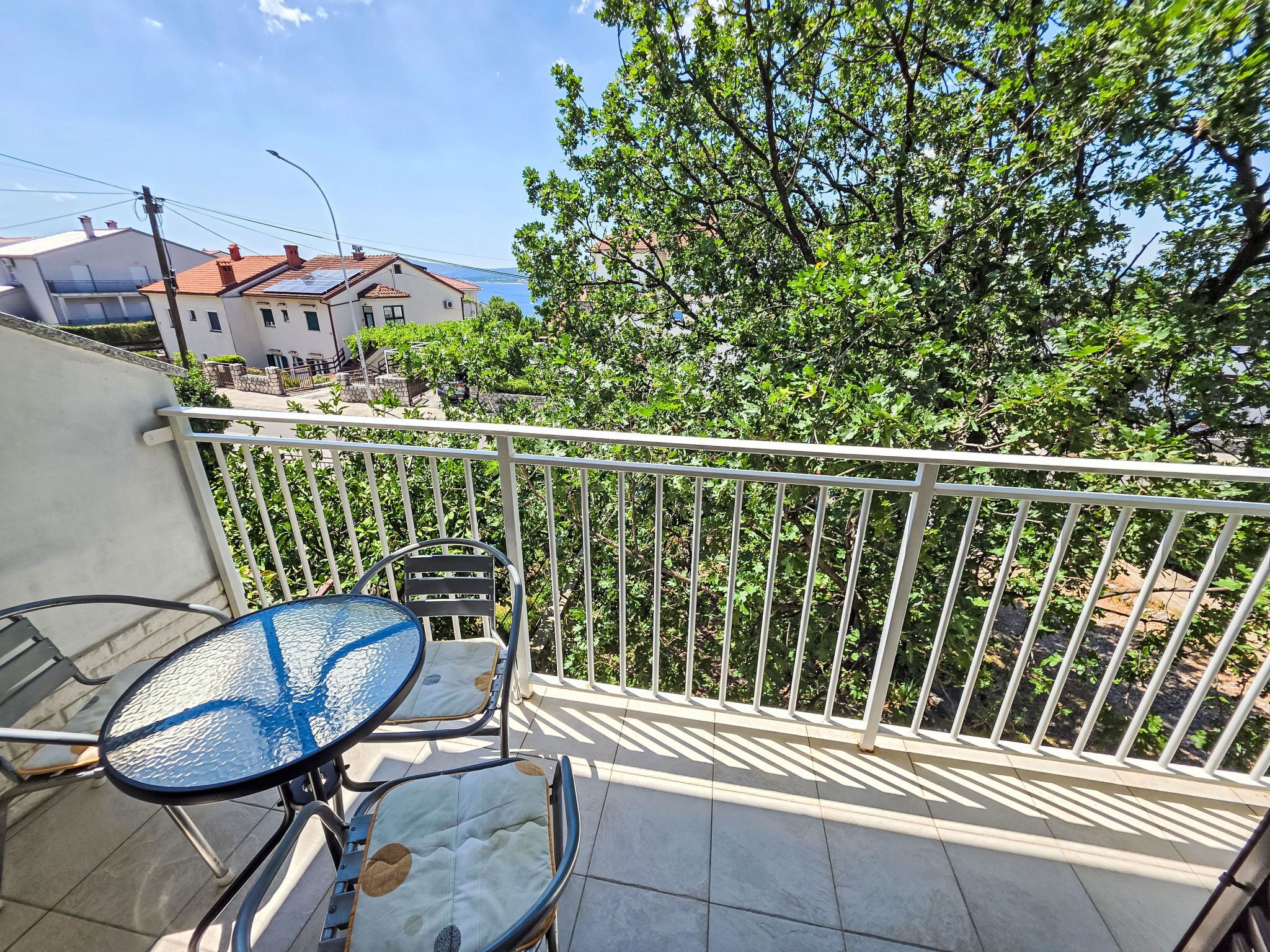 A balcony with a table and chairs, a white railing, and a view of trees and buildings.