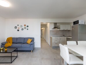 Eine moderne Wohnung mit Wohnbereich, ausgestattet mit einer Couch, einem Couchtisch und Wanddekoration. Eine Küche mit Kühlschrank, Mikrowelle und einem Essbereich mit Stühlen ist sichtbar.