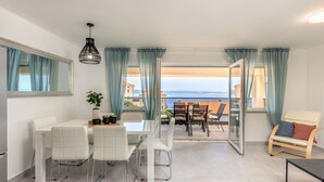 Ein modernes Wohnzimmer mit Essbereich, großen Fenstern mit Vorhängen, Meerblick und einem Balkon mit Gartenmöbeln.
