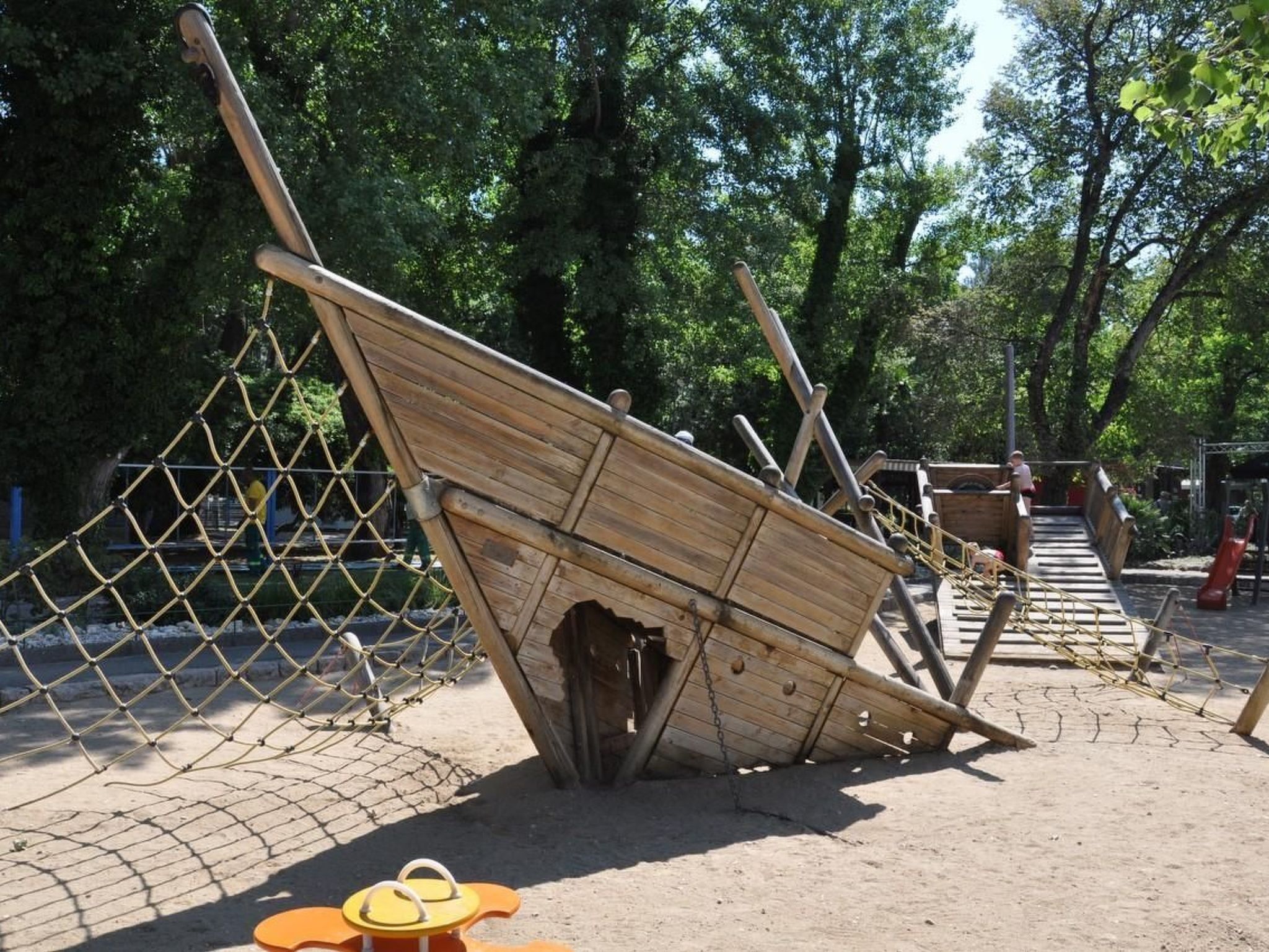 Estructura de un naufragio de madera en un parque, con una cerca de alambre y un parque infantil al fondo.