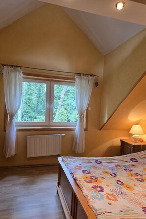 A bedroom with a sloped ceiling, a bed with a floral bedspread, a window with curtains, a radiator, and a bedside lamp. - Charmantes Haus 100m vom Strand entfernt für 6 Personen by Interhome (Pobierowo)