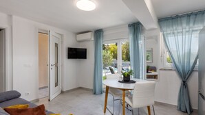 Ein modernes Wohnzimmer mit einer Couch, einem Esstisch mit Stühlen, einem an der Wand montierten Fernseher und Blick ins Freie durch große Fenster.