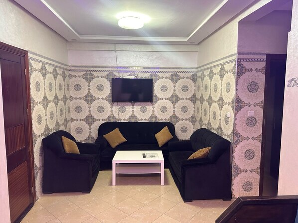 Un salón con rincón amueblado, con papel tapiz estampado, un sofá negro, dos sillones, una mesa de centro blanca y un televisor de pantalla plana montado en la pared.