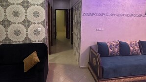 A living room with a black sofa and a blue sofa with patterned cushions, a patterned wall, and a hallway with a door leading to another room. - Residence Ennassr 3 by Interhome (Agadir)